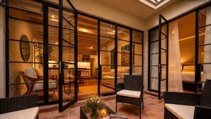 Amplia zona de patio con mobiliario de mimbre, una mesa de centro con un libro y flores, y vistas al interior a través de grandes puertas de vidrio.