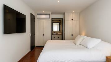 A bedroom with a large bed, a TV mounted on the wall, a built-in wardrobe, and a bathroom with a mirror and sink.
