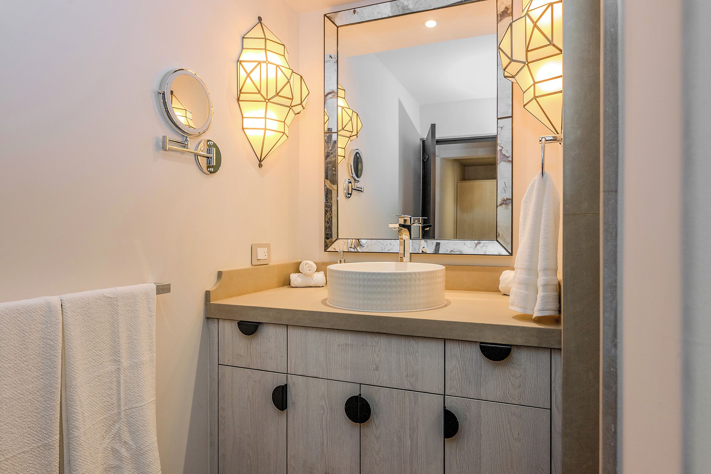 A bathroom with a round mirror, two wall-mounted lights, a round sink, and a towel rack.