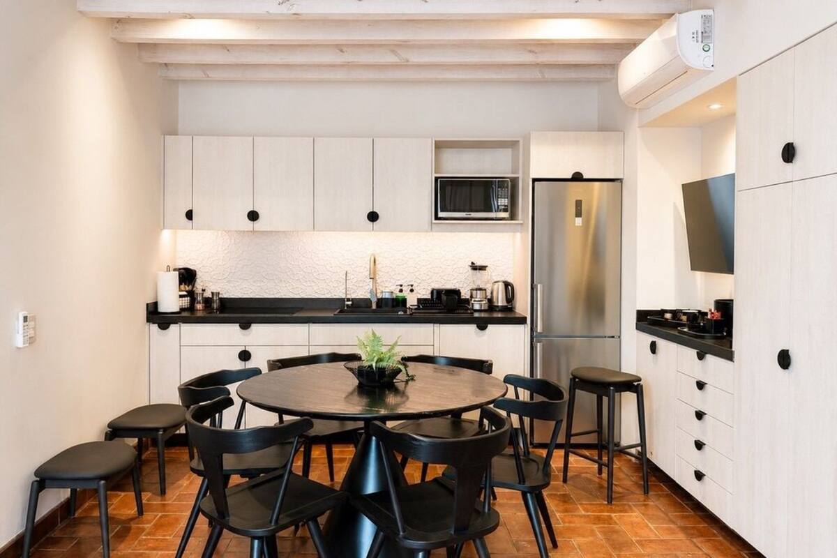 Une cuisine compacte avec des meubles blancs, une table de salle à manger ronde, des chaises noires, un four à micro-ondes et un réfrigérateur en acier inoxydable.