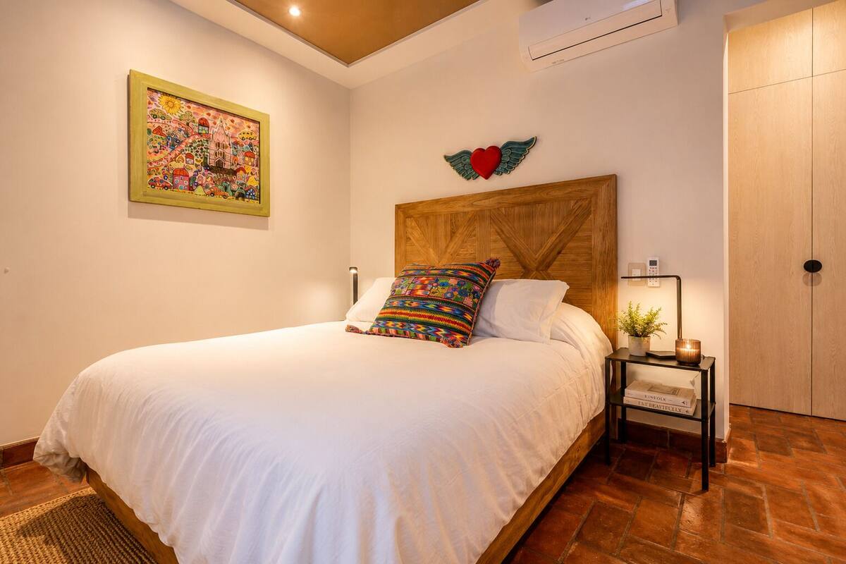 Un lit bien fait, avec du linge de lit blanc, un coussin coloré, une tête de lit en bois, un climatiseur fixé au mur, un tableau encadré et une table de chevet avec une lampe et des livres.