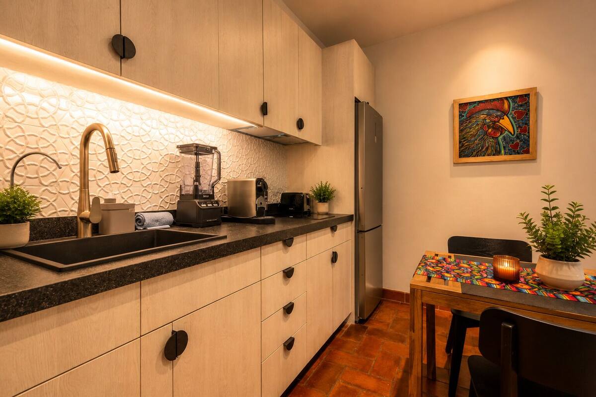 Une cuisine moderne équipée d’un évier intégré, d’un mixeur, d’une machine à café et d’un réfrigérateur. On y trouve une table à manger avec des chaises et un chemin de table coloré.
