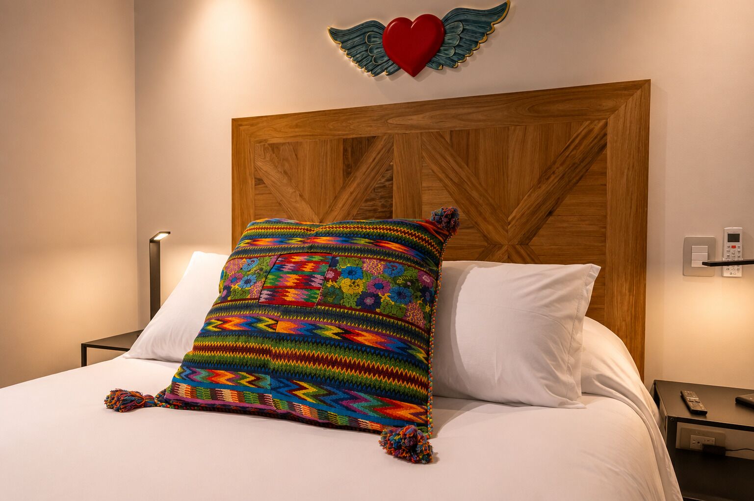 A bed with a colorful blanket, a wooden headboard, a wall-mounted heart decoration, and a bedside table with a remote control.