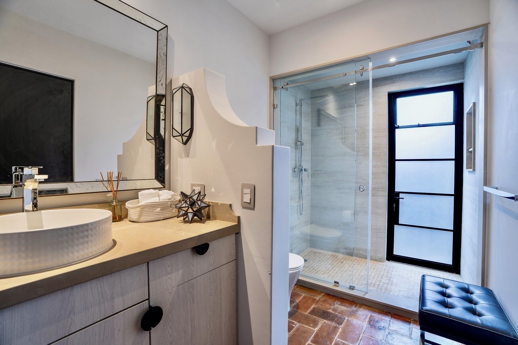 A modern bathroom with a large mirror, a round sink, a glass shower enclosure, and a black leather bench.
