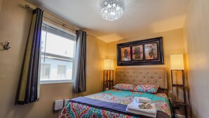 A hotel room with a bed, two bedside lamps, a chandelier, a window with curtains, and artwork on the wall. - Finding Your Rhythm In The Heart Of It All (Colorado Springs)