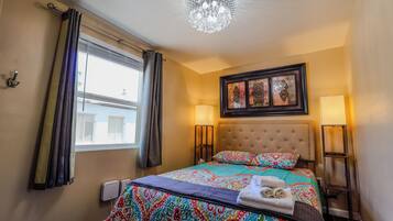 Une chambre d’hôtel avec un lit, deux lampes de chevet, un lustre, une fenêtre avec des rideaux et des œuvres d’art accrochées au mur.