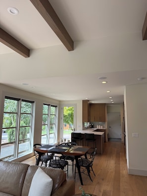 A spacious living area with a large window, a dining table with chairs, and a kitchen in the background. - Modern 2-bedroom with views on acreage w/sport court/pickle-ball on Fox Island (Fox Island)