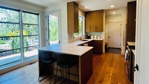 A modern kitchen with a large island, wooden flooring, and a view of greenery outside.