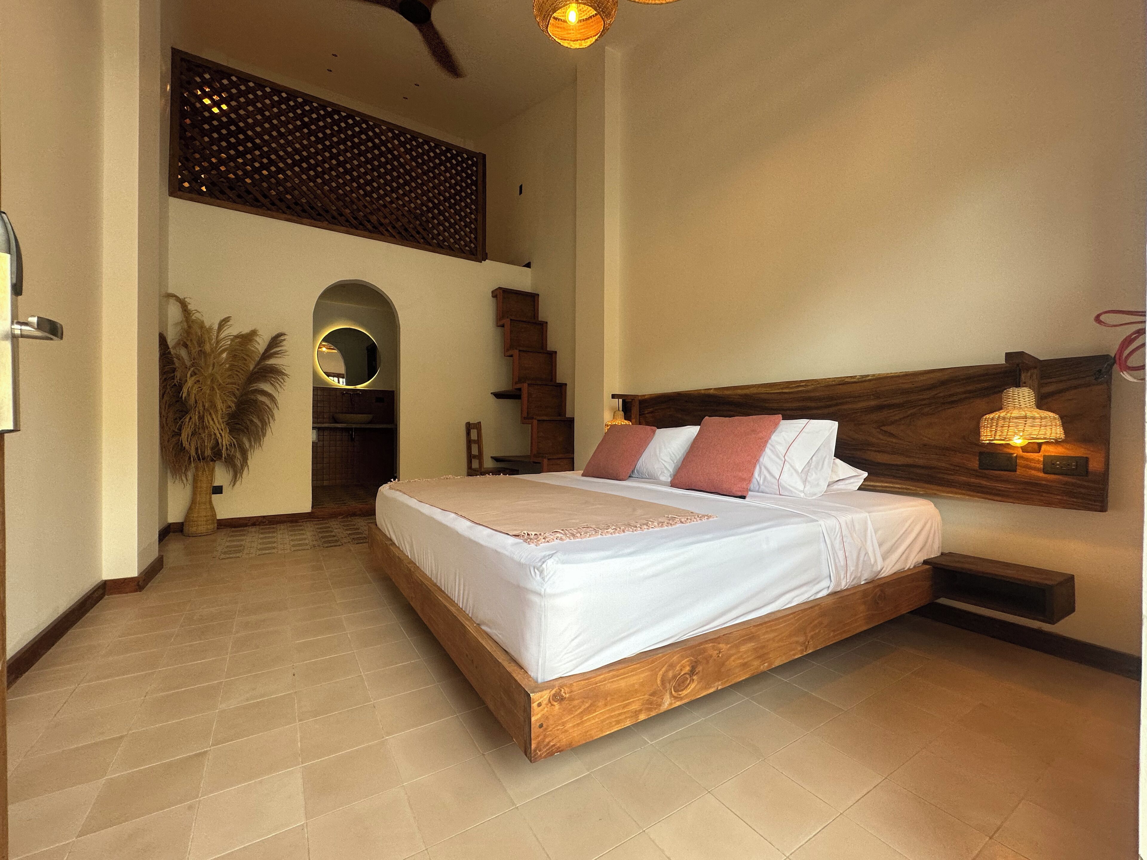 A bedroom with a large bed, a wooden headboard, a small table, a wall-mounted lamp, and a staircase leading to a bathroom.
