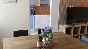 A modern living room with a wooden dining table, a TV on a stand, and a kitchen in the background. - Apartment near american school from the center of milan from IEO and Humanitas (Noverasco)