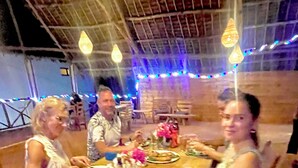 A group of people seated around a wooden table with plates of food, under a tent-like structure with hanging lights. - Zanzibar beach escape  (Bwejuu)