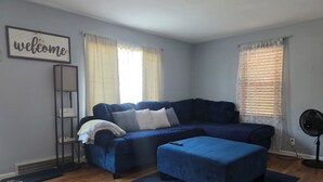 A living room with a dark blue sofa, an ottoman, a standing fan, and a 'welcome' sign on the wall. - Charming Cape Cod minutes from YSU, Covelli Center and many more! Pet Friendly (Youngstown)