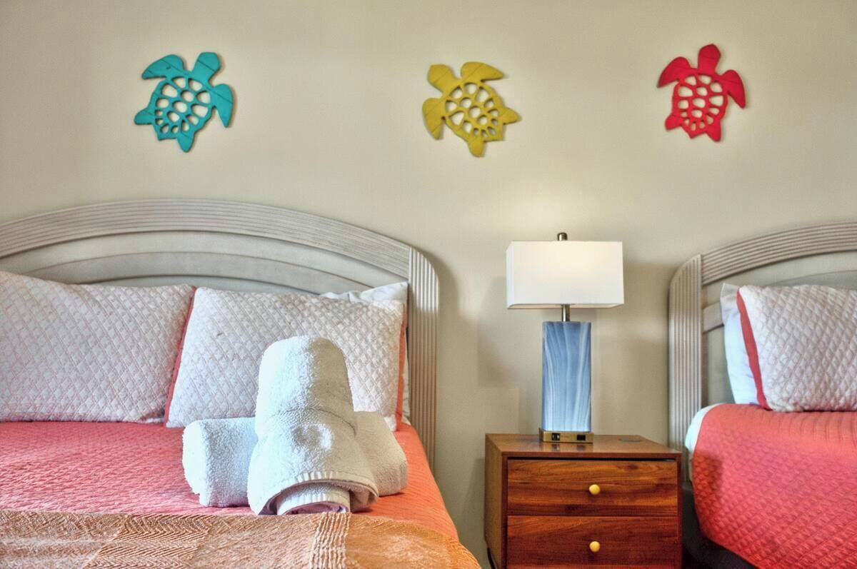 A hotel room with two beds, each with a headboard, a bedside table with a lamp, and decorative turtle wall art.