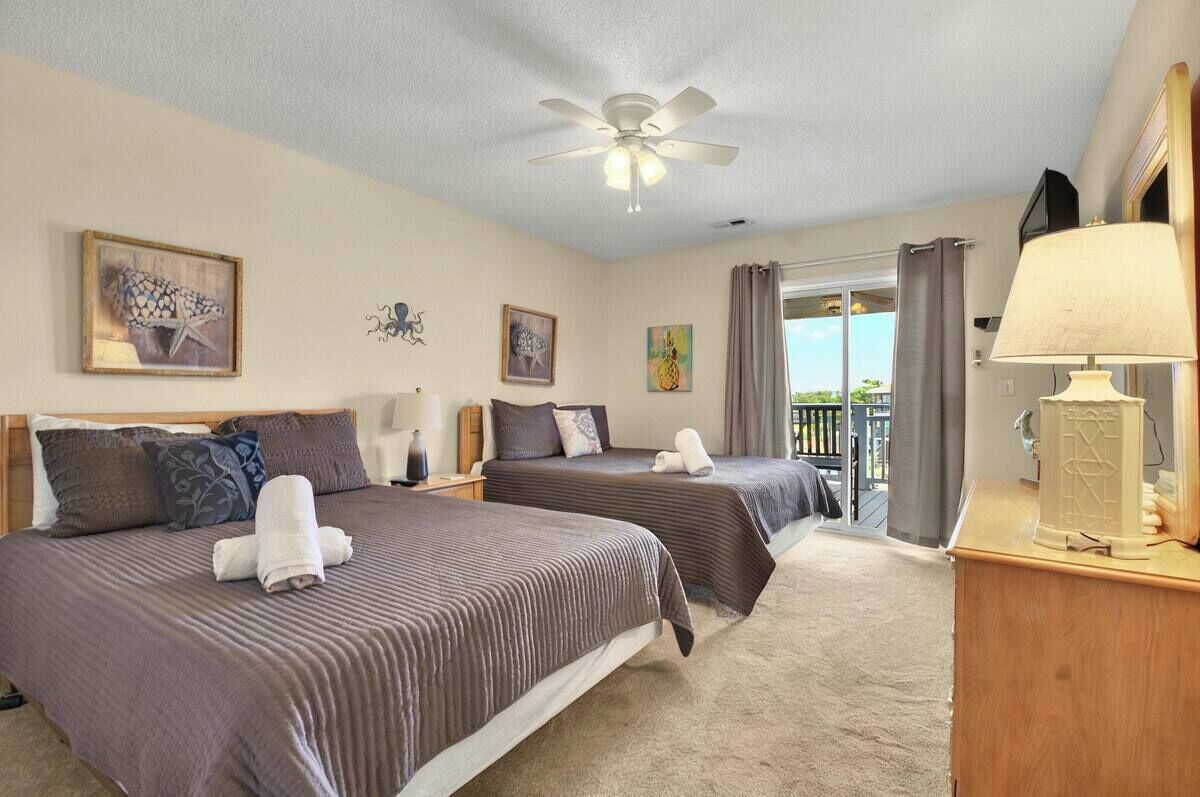 A hotel room with two beds, a ceiling fan, artwork on the walls, a window with curtains, and a lamp on a bedside table.