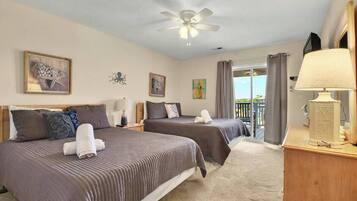 A hotel room with two beds, a ceiling fan, artwork on the walls, a window with curtains, and a lamp on a bedside table.