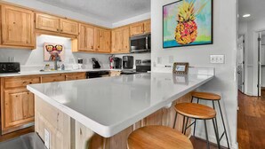 Une cuisine moderne avec des meubles en bois, un grand îlot central doté de tabourets et un tableau coloré représentant un ananas accroché au mur.