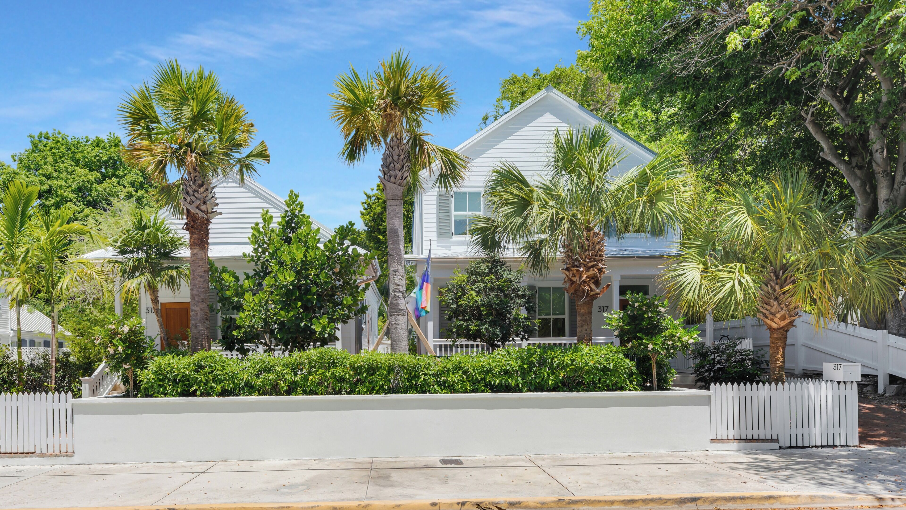 A two-story white house with a white picket fence, surrounded by lush greenery and palm trees.