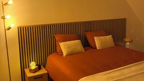 Une chambre avec un plafond en pente, un lit doté d’une tête de lit en bois, une table de chevet avec une lampe et un lampadaire à plusieurs ampoules.
