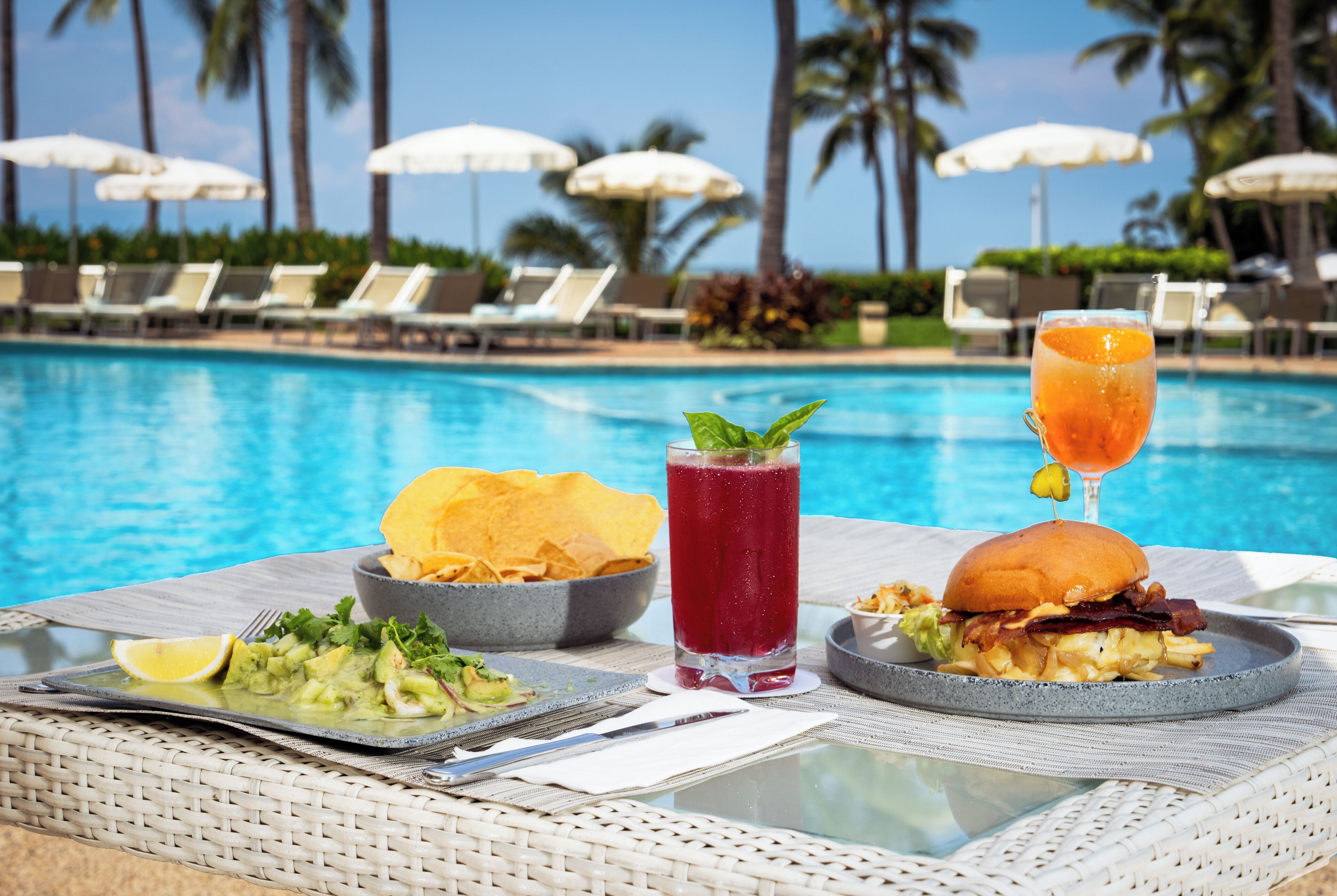 Une table à manger avec une assiette de nourriture, une boisson et un sandwich, placée près d’une piscine.