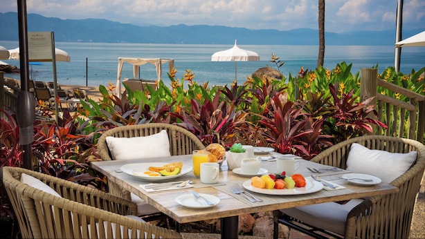 A dining table set with plates of fruit, a croissant, and a cup of coffee, overlooking a body of water with mountains in the distance. - Ocean Breeze Puerto Vallarta (Puerto Vallarta)