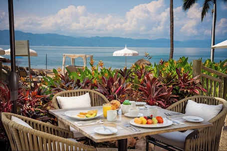 A dining table set with plates of fruit, a croissant, and a cup of coffee, overlooking a body of water with mountains in the distance.