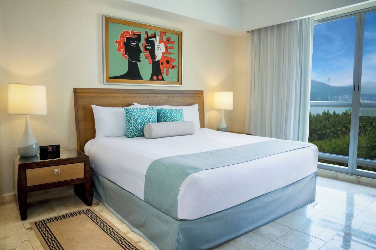 Une chambre d’hôtel avec un grand lit, deux lampes de chevet, une horloge numérique, une œuvre d’art encadrée au-dessus du lit et une vue sur l’extérieur grâce à une grande fenêtre.