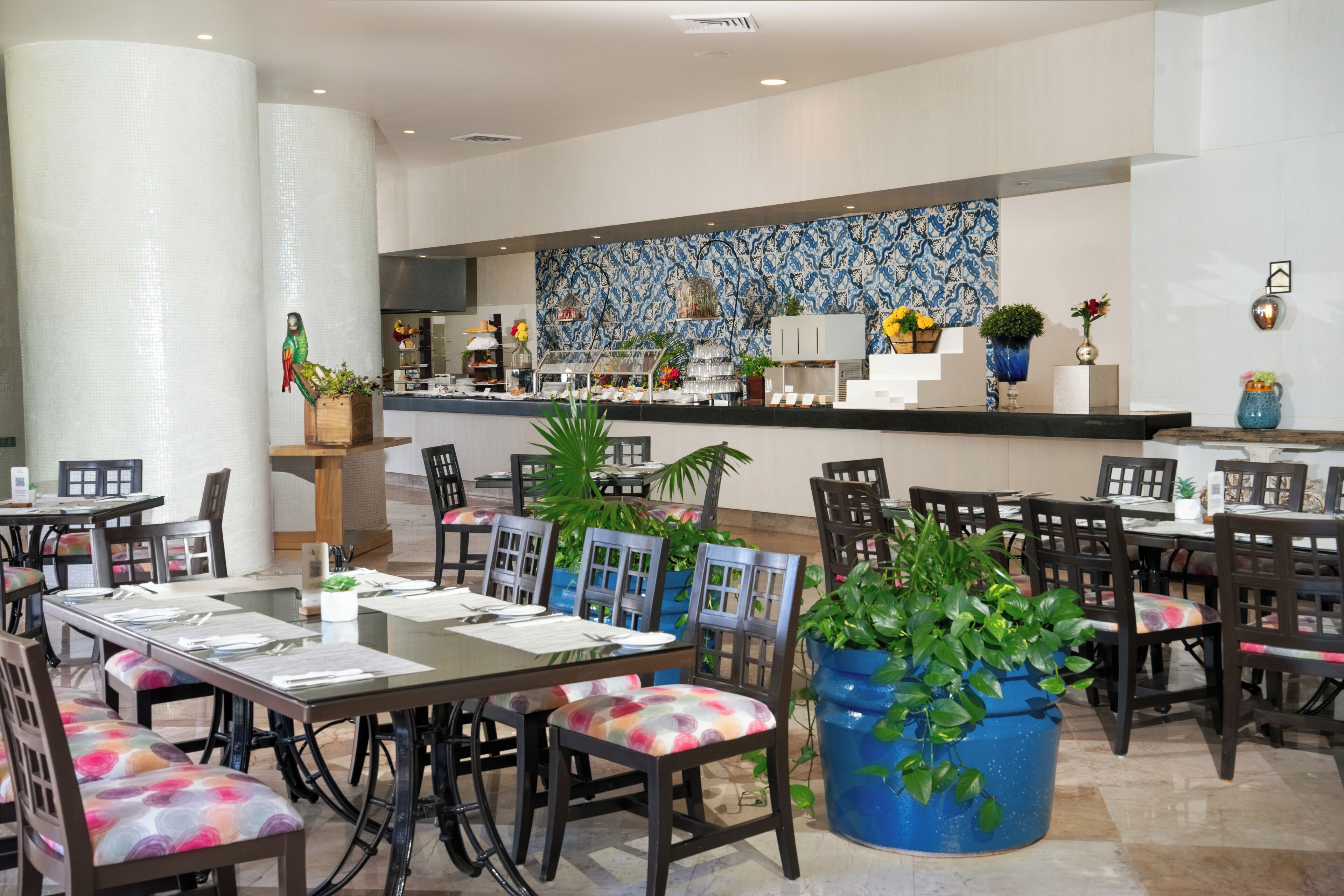 Een restaurant met meerdere tafels en stoelen, een toonbank met een blauwe, geruite spatwand en diverse planten en decoraties.