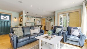 Ein Wohnzimmer mit einem dunkelblauen Sofa, einem weißen Couchtisch und einem blauen Sessel. Im Hintergrund befindet sich eine Küche mit weißen Schränken und einer Holztür.