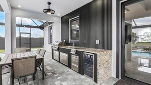 A modern outdoor kitchen with a dining area, built-in appliances, and a view of a swimming pool.