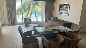 Un'ampia zona giorno con tavolo da pranzo in vetro circondato da sedie in vimini, un divano e una grande finestra con vista su palme e sull'oceano.