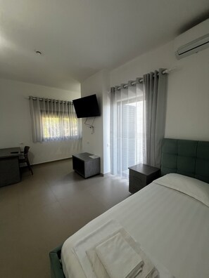 A hotel room with a bed, a desk, a chair, a TV mounted on the wall, and a window with curtains.