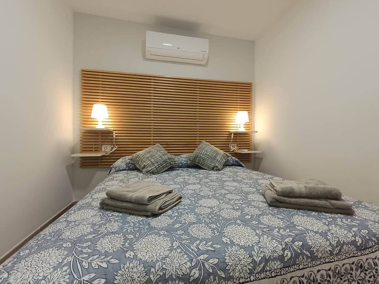 Un lit bien fait, recouvert d’une couvre-lit à motifs floraux, avec deux lampes de chevet et un climatiseur fixé au mur.
