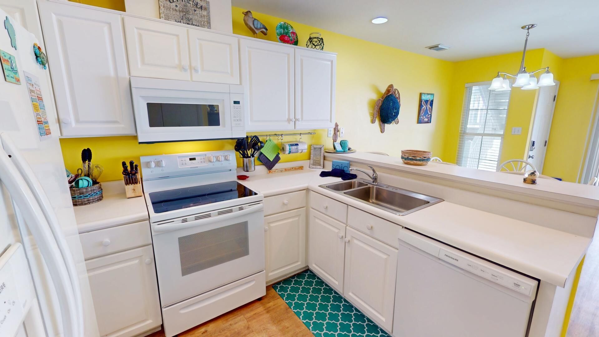 Una cocina compacta con muebles blancos, microondas, estufa y lavavajillas. Presenta una pared amarilla, piso estampado y varios utensilios y adornos de cocina.