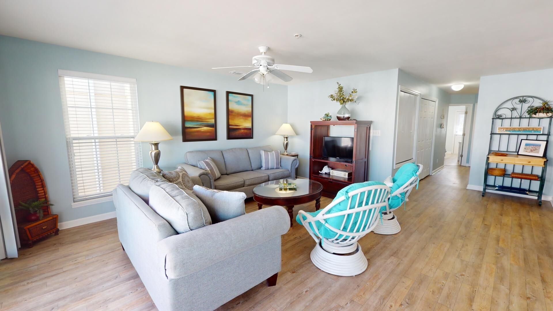 A living room with a grey sofa, a coffee table, and two turquoise chairs. There is a ceiling fan, a TV on a wooden cabinet, and a bookshelf with decorative items.