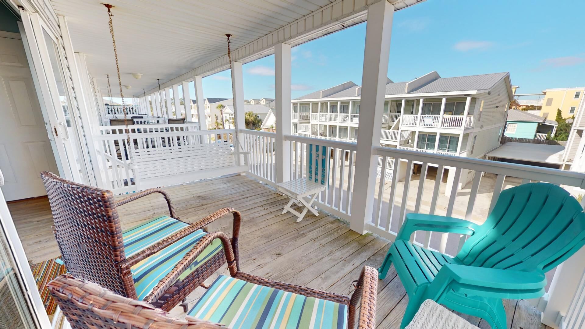 A covered porch with wicker chairs and a turquoise chair, overlooking a row of white buildings.