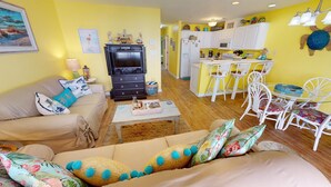 A living room with a sofa, coffee table, and a TV. There is a kitchen area with white cabinets and a dining table with chairs. - South Beach Ocean S by Tybee Vacation Rentals (Tybee Island)