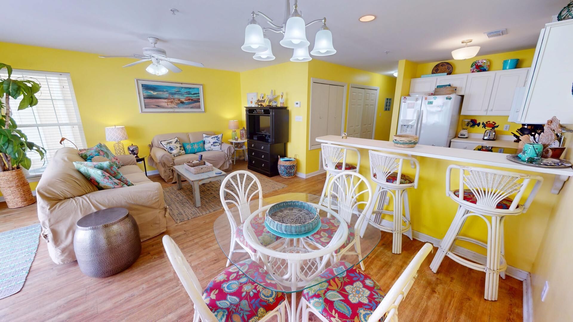 A bright yellow living room with a sofa, a dining table with chairs, a kitchen area with a fridge, and a ceiling fan.
