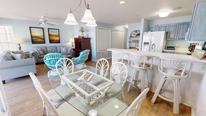 A modern living room with a glass dining table, white wicker chairs, a sofa, and a ceiling fan. - South Beach Ocean S by Tybee Vacation Rentals (Tybee Island)