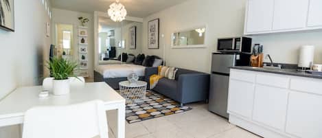 A compact living space with a dining area, a gray sofa with cushions, a kitchen with a microwave and refrigerator, and a patterned rug.