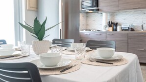 Un comedor con una mesa lista para cuatro personas, que incluye platos, tazones y vasos blancos. Hay una planta en maceta sobre la mesa, y se observan muebles de madera y una cocina moderna al fondo.