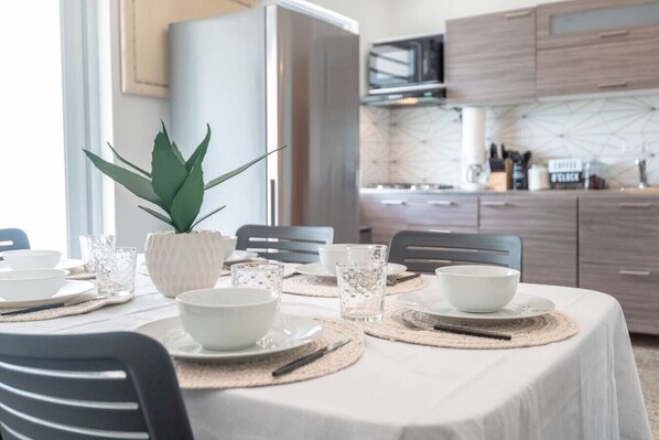 A dining area with a table set for four, including white plates, bowls, and glasses. There is a potted plant on the table, and the room has wooden cabinets and a modern kitchen in the background.
