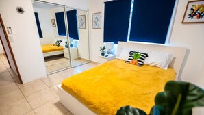 A bedroom with a bed, a nightstand, a mirror, and a window with blue curtains. - Casa Paloma「UNIT 2 「Judith」Walk to beach (San Juan)