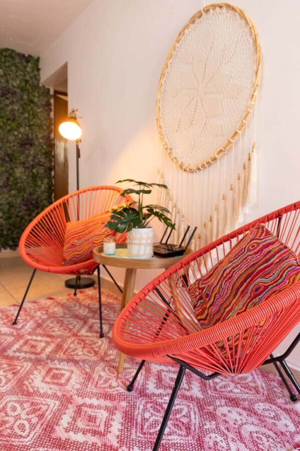 A room with two orange woven chairs, a round table with a plant, and a large wall hanging.