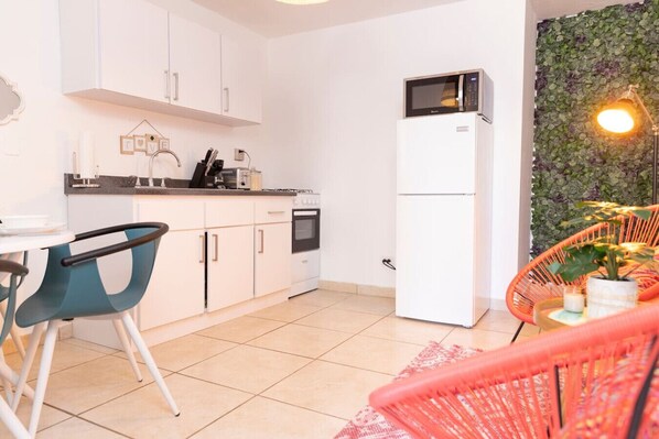 A compact kitchen with white cabinets, a microwave, and a refrigerator. There is a dining area with a blue chair and a small table.