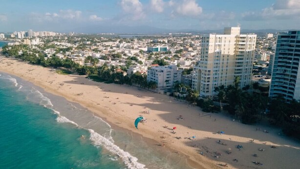 An aerial view of a beach with a kite surfer, beachfront buildings, and a clear sky. - Casa Paloma「UNIT 2 「Judith」Walk to beach (San Juan)