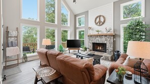 A spacious living room with a stone fireplace, large windows, and a television.