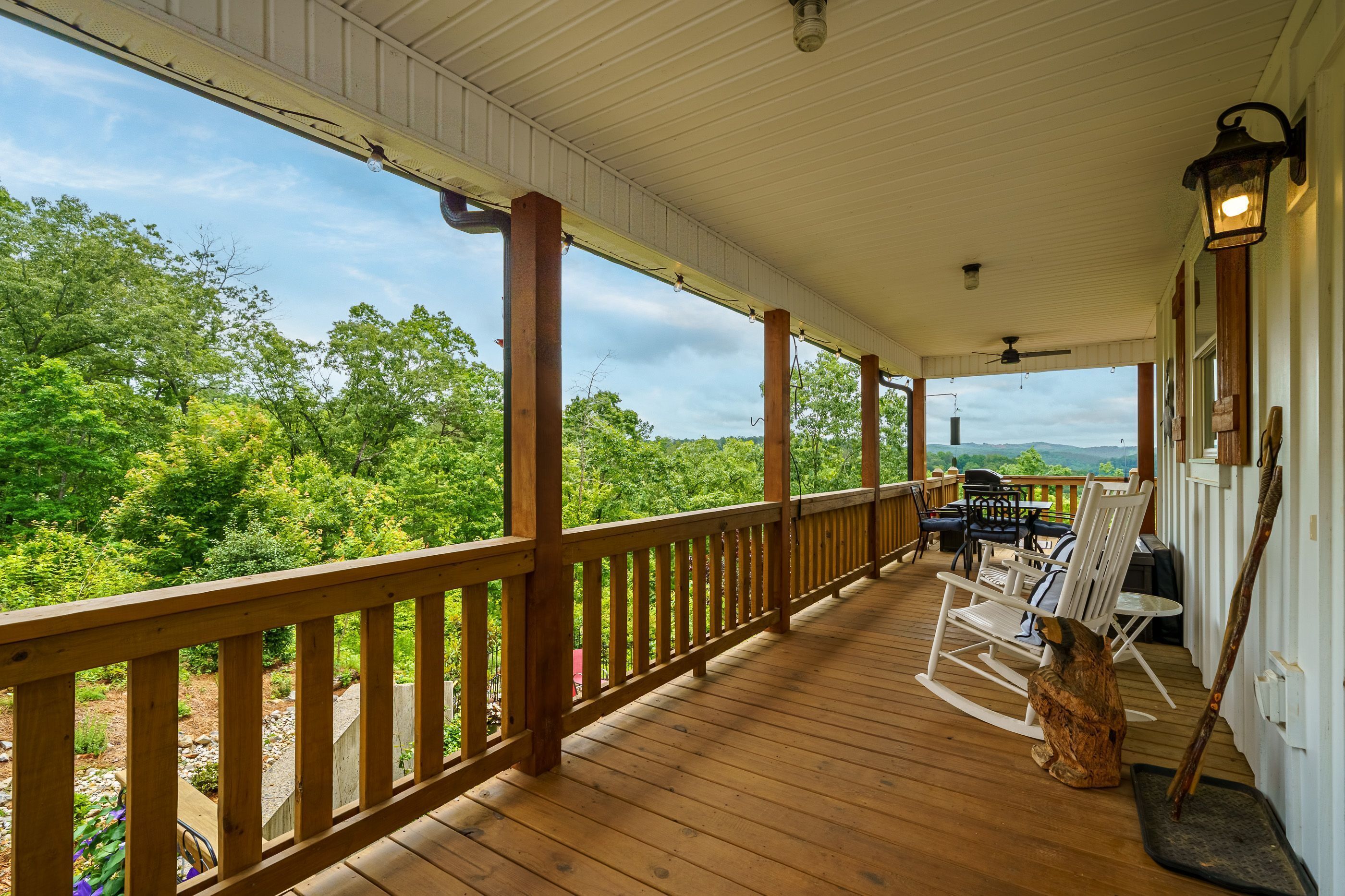 A covered porch with a wooden railing, outdoor furniture, and a view of a forested landscape.