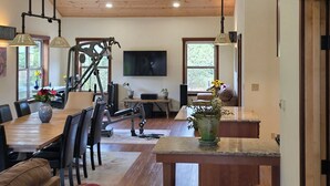 A spacious living area with a wooden ceiling, a ceiling fan, a dining table with chairs, a TV mounted on the wall, and a large window providing natural light.