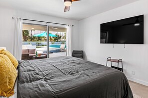 Ein Schlafzimmer mit einem Bett, einem an der Wand montierten Fernseher und einer Glastür, die zu einem Außenbereich mit Pool und Sonnenschirmen führt.