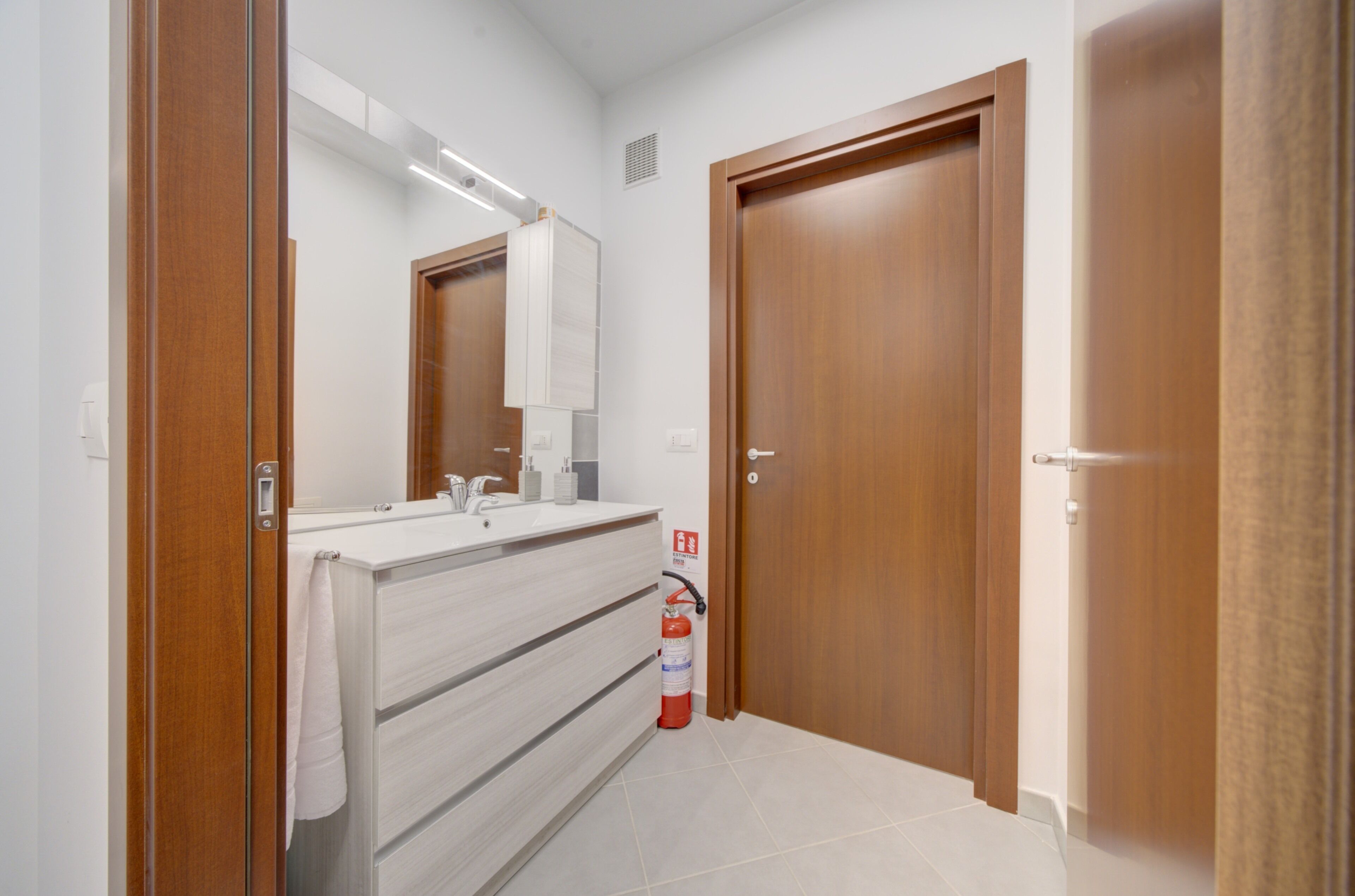 A modern bathroom with a wooden vanity, a mirror, and a fire extinguisher.